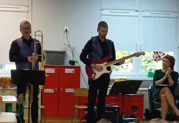 Pani siedzi na krześle. jeden pan stoi i gra na gitarze elektrycznej, drugi pan trzyma w ręku puzon.