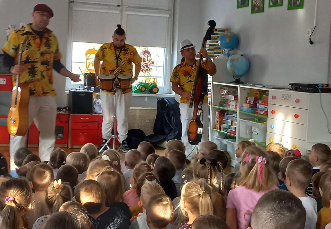 Trzech panów gra na instrumentach: saksofon, gitara, bongosy. Dzieci siedzą na dywanie.