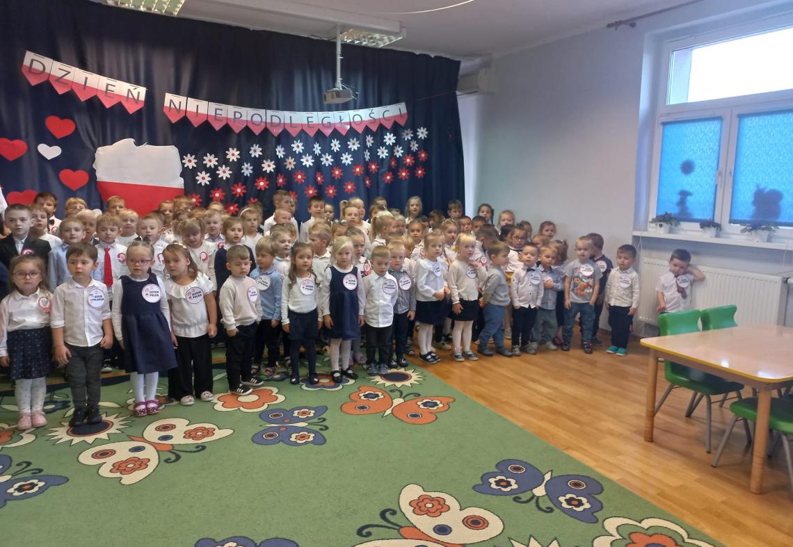 Dzieci z wszystkich grup stoją na tle dekoracji o Polsce i śpiewają hymn.