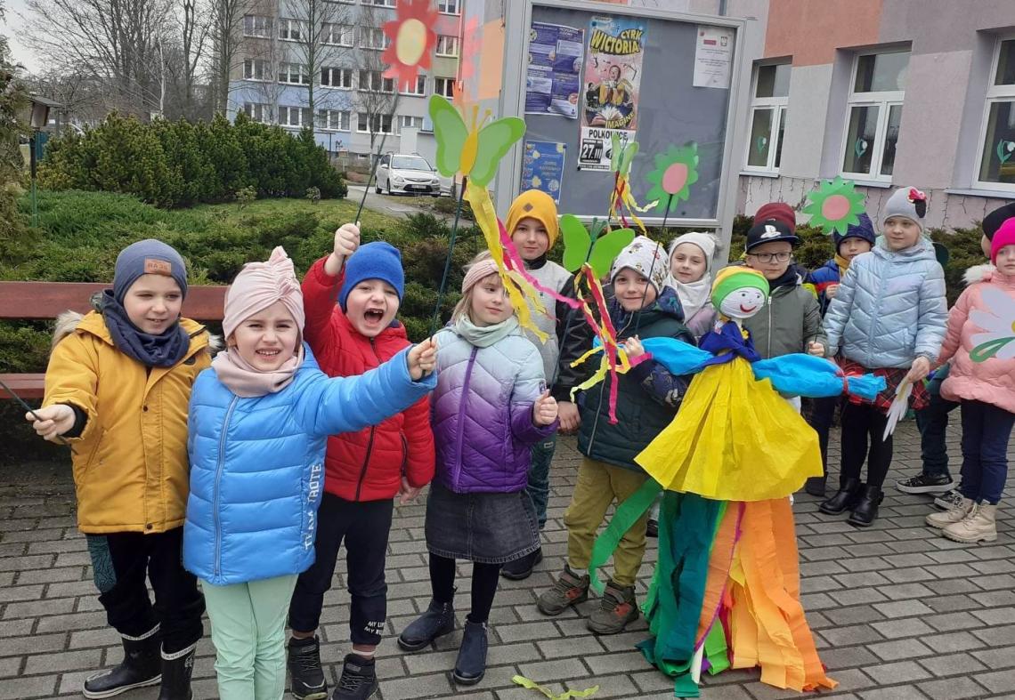 Dzieci stoją przed przedszkolem i witają wiosnę machając korowymi kwiatami
