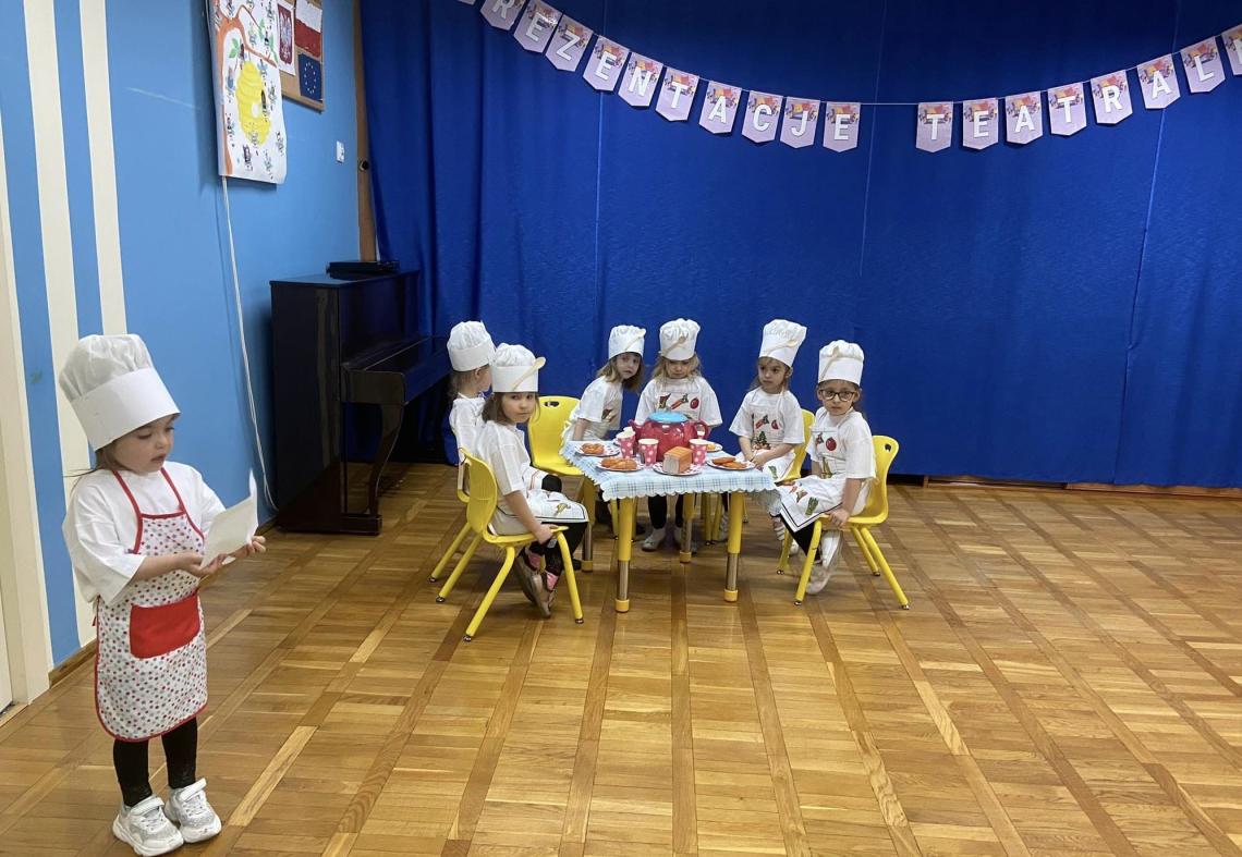 Sześć dziewczynek przebranych za kucharki siedzi przy stoliku. Jedna dziewczynka stoi 