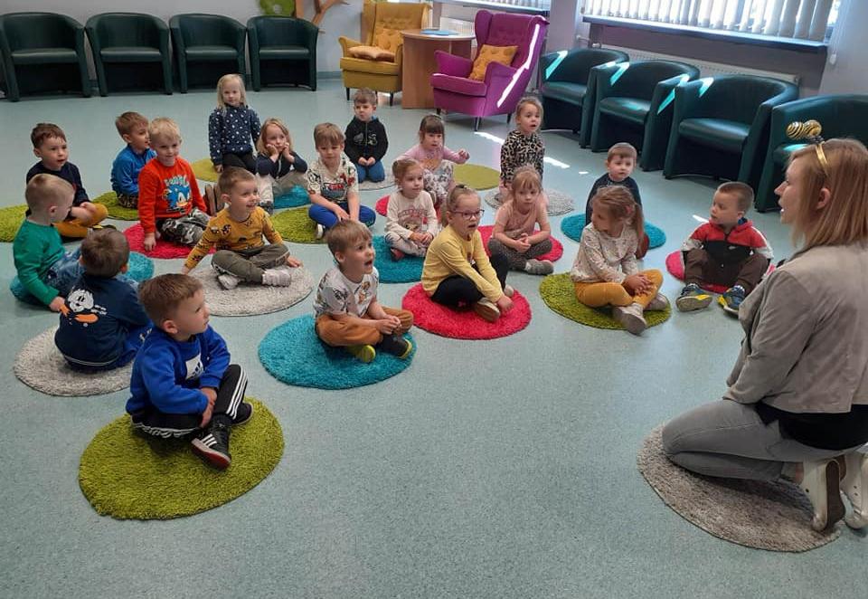 Dzieci siedzą na małych dywanikach na zajęciach w bibliotece.