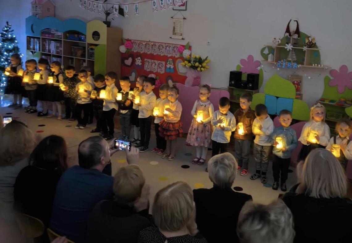 Dzieci stoją w półkolu trzymając w ręku zapalone lampki.