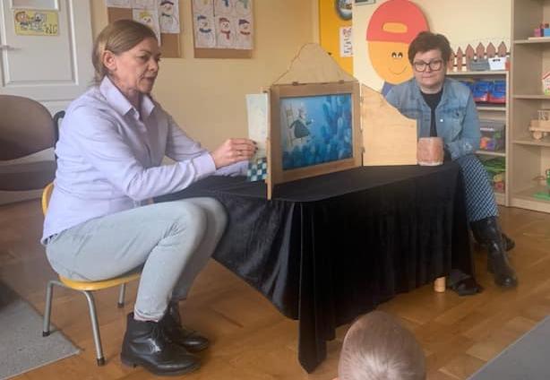 Dwie panie siedzą na krzesełkach i przedstawiają teatrzyk Kamishibai.