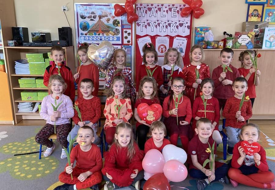Dzieci ubrane na czerwono stoją w grupie trzymając w ręku kwiatka lub serce.