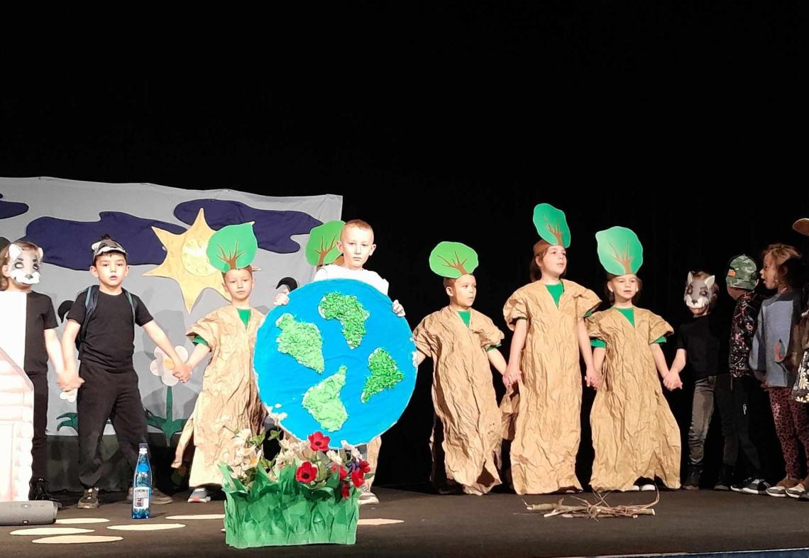 Dzieci przebrane za drzewa i zwierzęta stoją na scenie.
