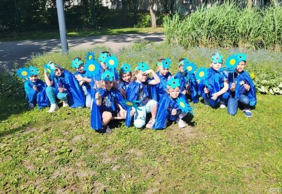 Dzieci ubrane w niebieskie pelerynki z opaskami kwiatowymi  na głowie kucają na trawie.