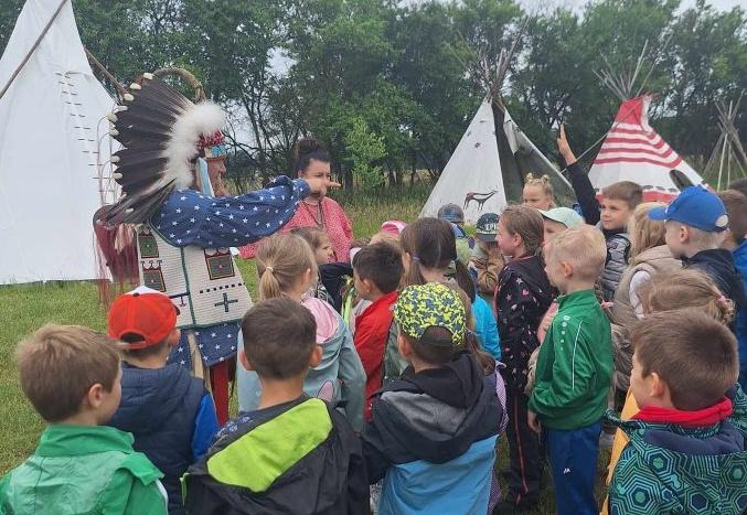 Dzieci stoją przy namiotach indiańskich i słuchają indianina.