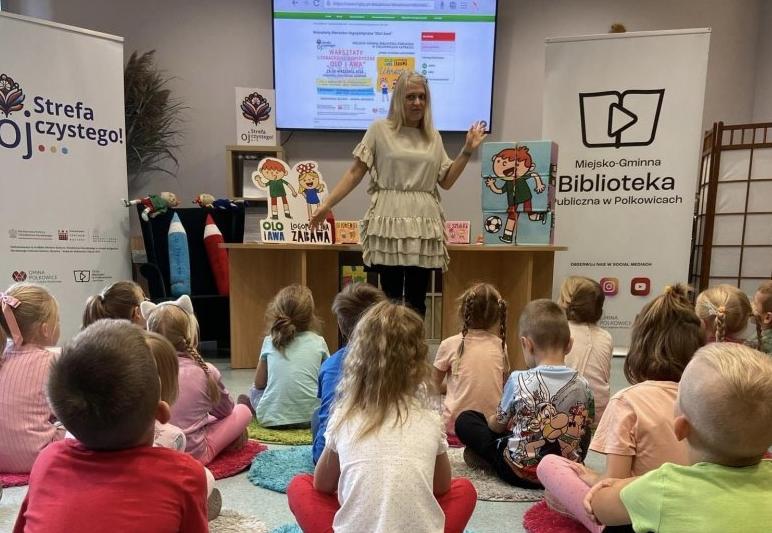 Dzieci siedzą na kolorowych dywanikach na zajęciach w bibliotece.