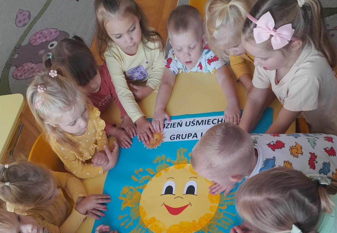 Dzieci ubrane na żółto stoją wokół plakatu ze słońcem i odciśniętymi rękami.