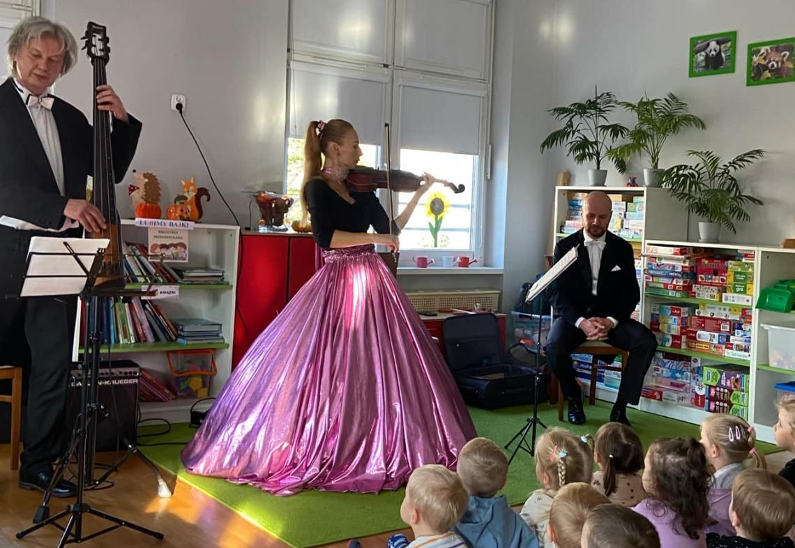 Troje muzyków gra na instrumentach muzycznych dzieci siedzą na dywanie i słuchają.