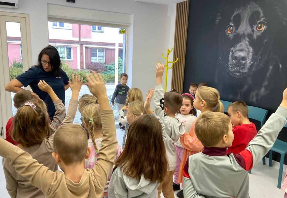 Dzieci  stoją w grupie słuchając pani weterynarz. Na ścianie obraz psa.