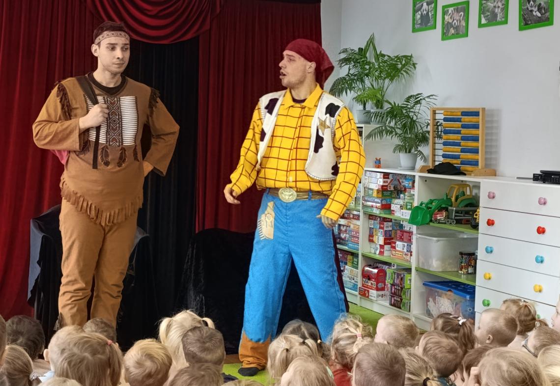 Dzieci siedzą na dywanie i oglądają teatrzyk w wykonaniu kowboja i kota.