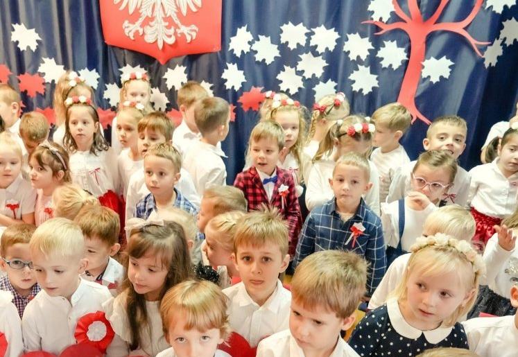 Dzieci ubrane na galowo trzymają w ręku czerwone serca z papieru i śpiewają.