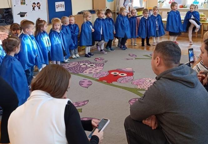 Dzieci w niebieskich pelerynkach stoją w półkolu. Rodzice oglądają występ.