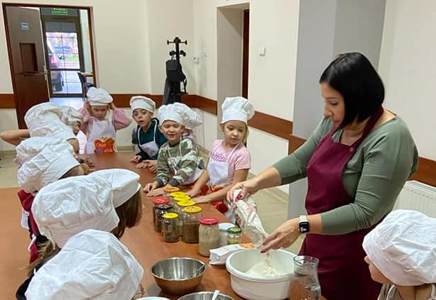 Dzieci przebrane za kucharzy stoją wokół stołu słuchając przepisu na chleb, który mówi pani.