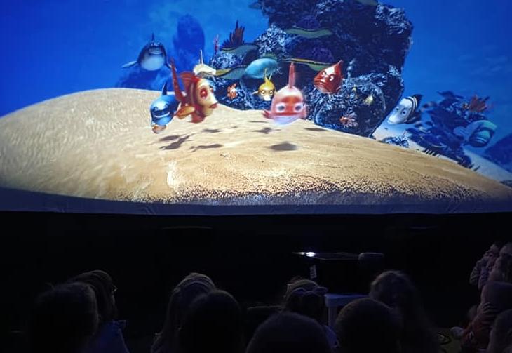 Dzieci siedzą pod kopułą i oglądają film o rybkach.