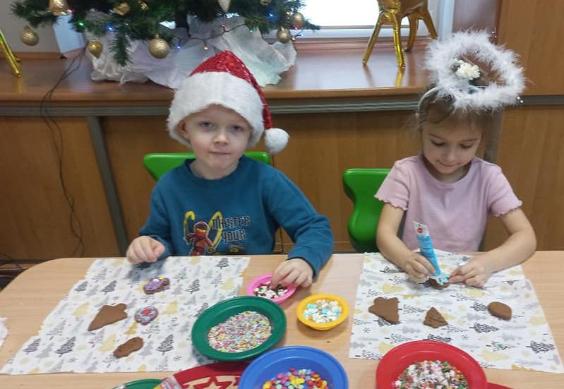 Chłopiec w mikołajkowej czapce i dziewczynka w aureoli siedzą przy stole i dekorują świąteczne pierniki.