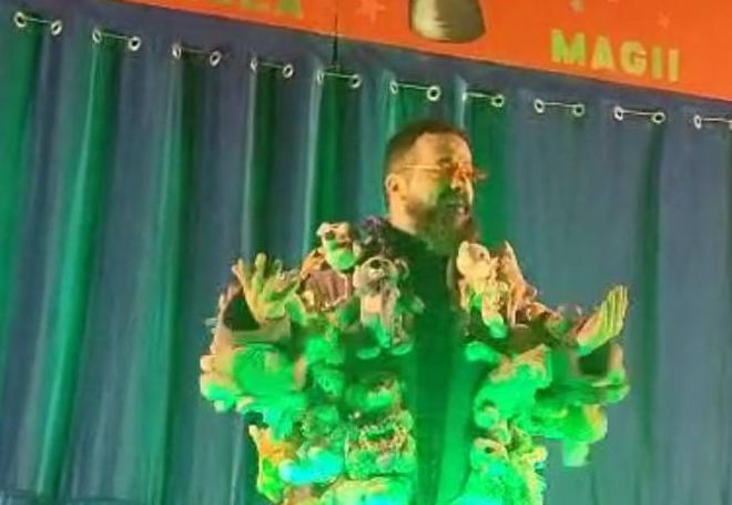 Na scenie stoi czarodziej ubrany w płaszcz ozdobiony maskotkami.