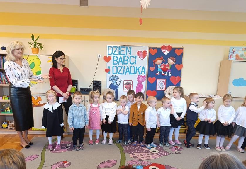 Dzieci ubrane na galowo stoją w półkolu razem z dwoma paniami.