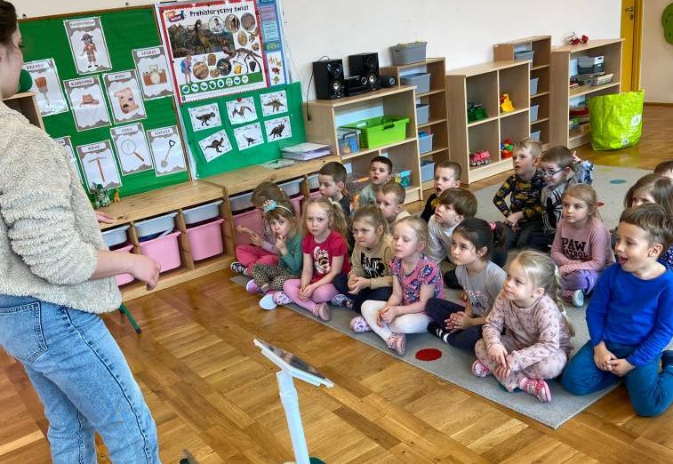 Dzieci siedzą w grupie na dywanie i słuchają pani, która opowiada o  energii odnawialnej.j.i 