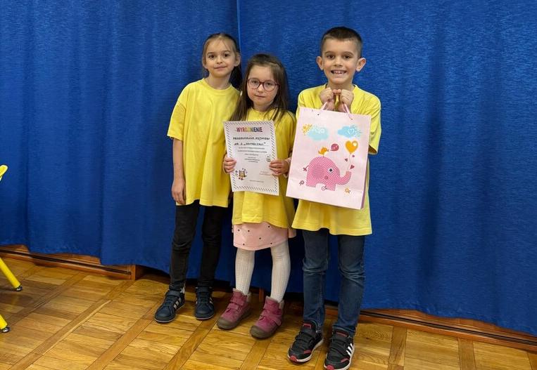 Troje dzieci stoi z dyplomem na tle napisu olimpiada matematyczna.