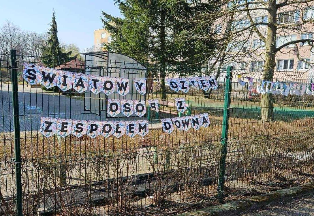 Skarpetki wykonane z papieru wiszą na sznurku na płocie przedszkola oraz napis światowy dzień osób z zespołem downa..