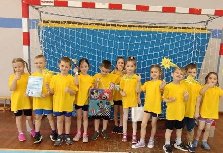 Grupa dzieci ubrana na sportowo w żółte bluzki stoi w bramce trzymając dyplom i puchar.