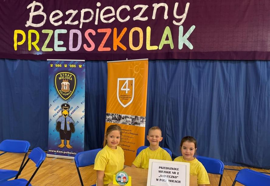 Dzieci ubrane w żółte koszulki siedzą przy stoliku na tle napisu bezpieczny przedszkolak.