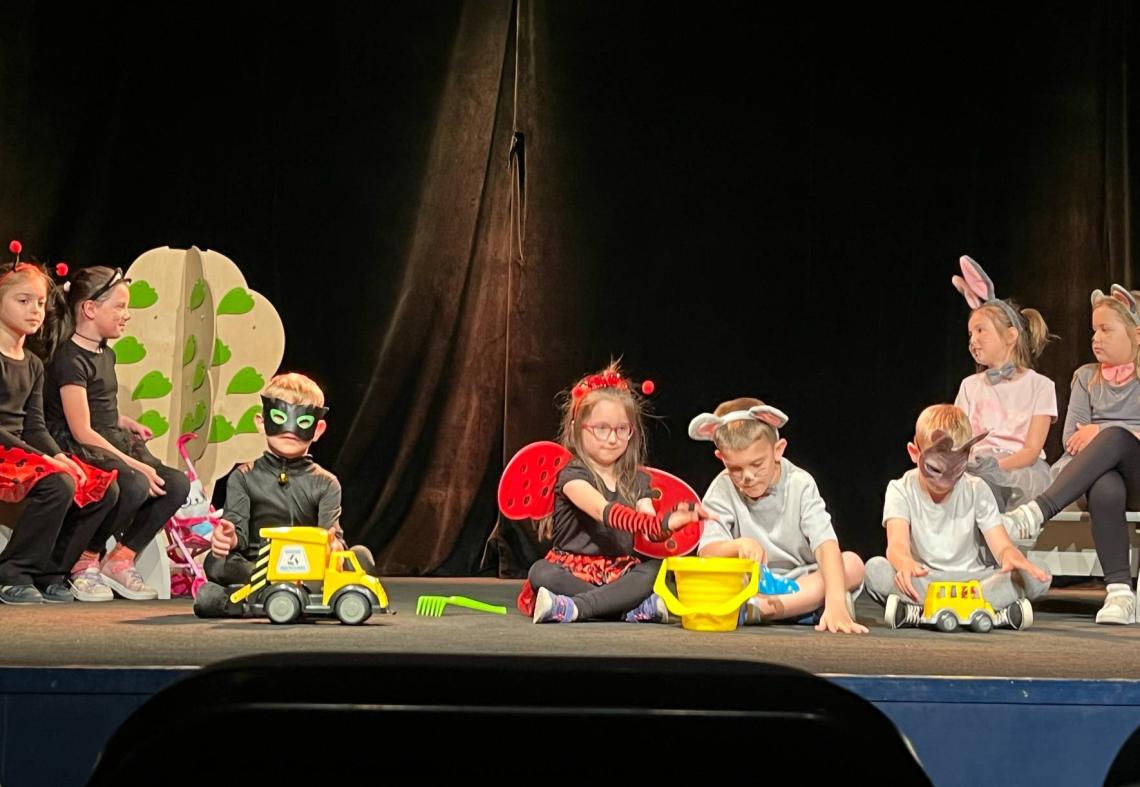 Dzieci przebrane za koty i biedronki oraz bawiące się samochodami występują na scenie.