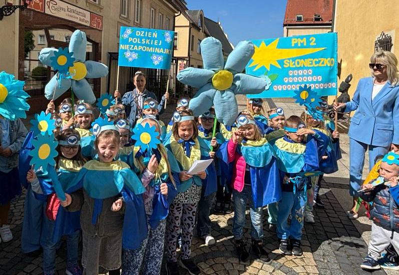 Dzieci ubrane na niebisko trzymają w ręku niebieskie kwiaty oraz plakaty.