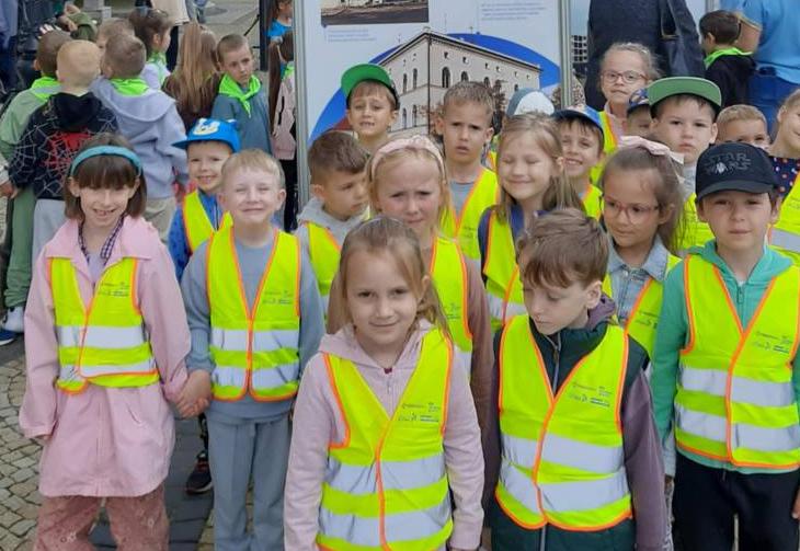 Dzieci stoja w grupie w kamizelkach odblaskowych na tle wystawy przedstawiającej Polkowice.