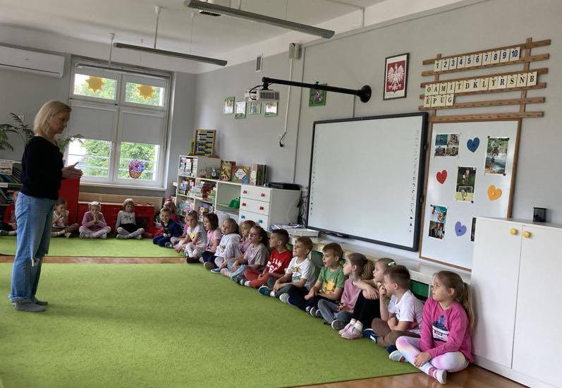 Dzieci siedzą w półkolu na dywanie razem z aktorką.