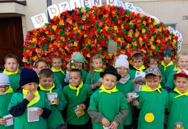 Dzieci ubrane na zielono stoją na tle kwiatów.
