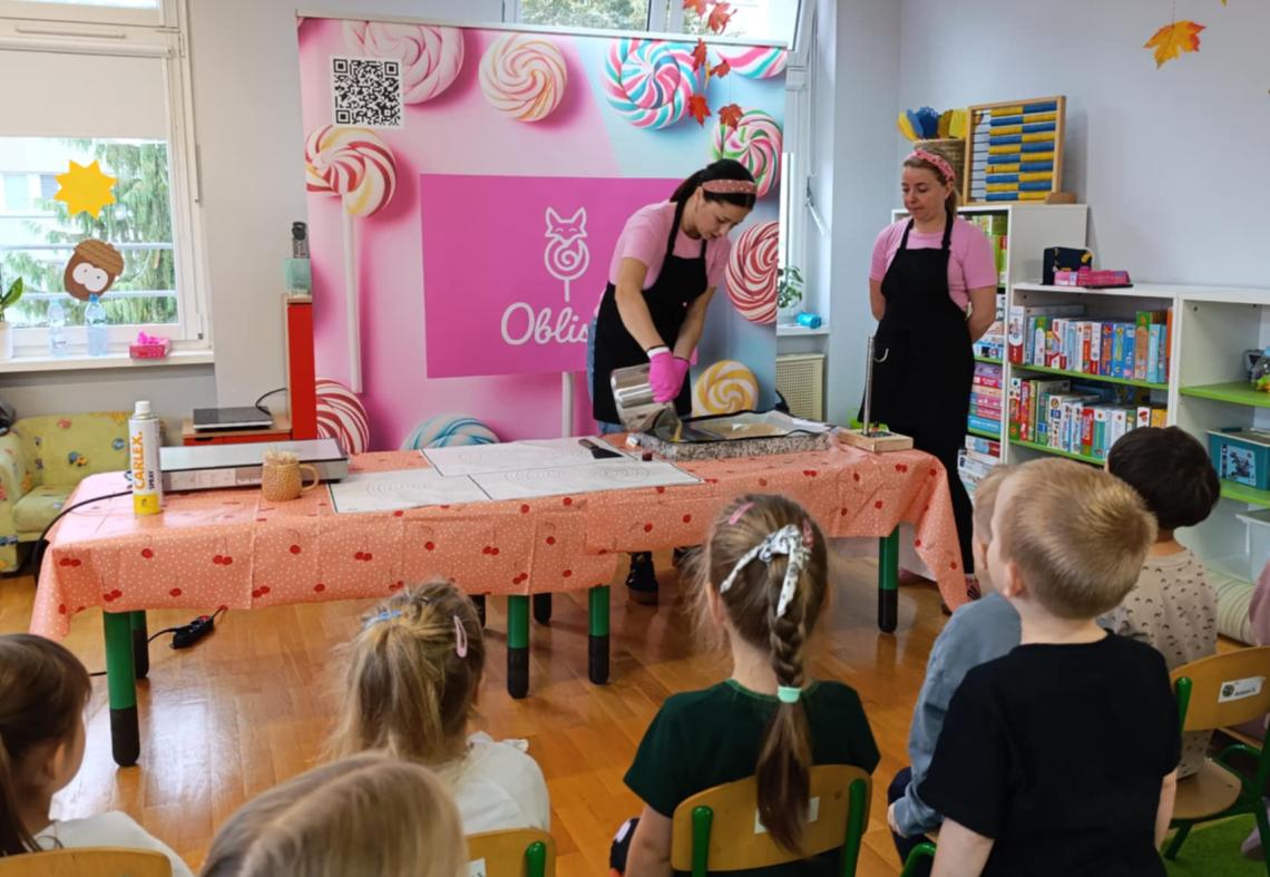 Dzieci siedzą na krzesełkach obserwują jak panie tworzą lizaki
