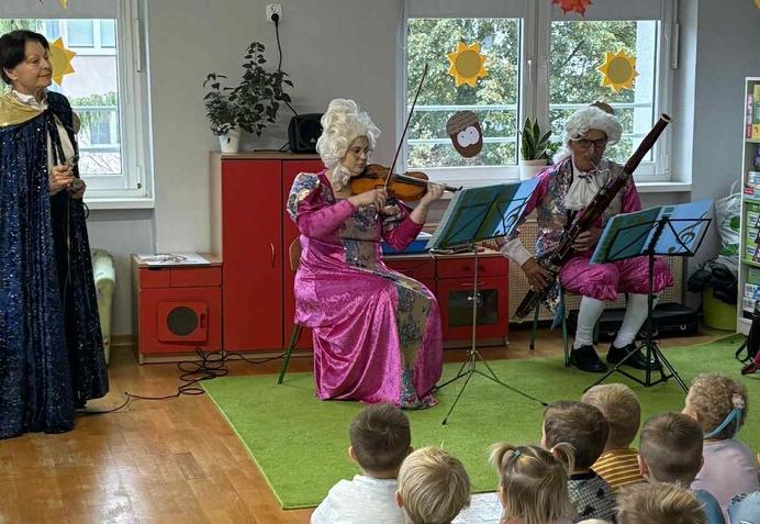 Dzieci siedząc na dywanie słuchają koncertu w  wykonaniu trzech muzyków