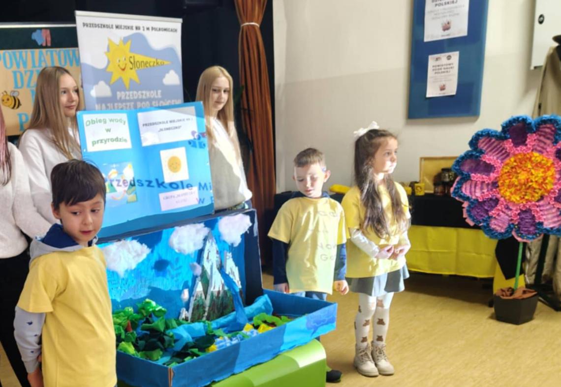 Troje dzieci ubranych w żółte koszulki stoi na tle makiety z obiegiem wody.