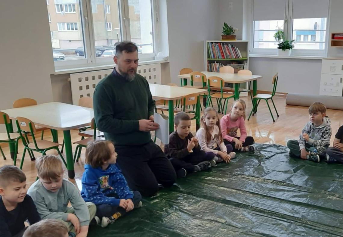 Dzieci siedzą w kole na dywanie przykrytym folią i słuchają zaproszonego gościa.