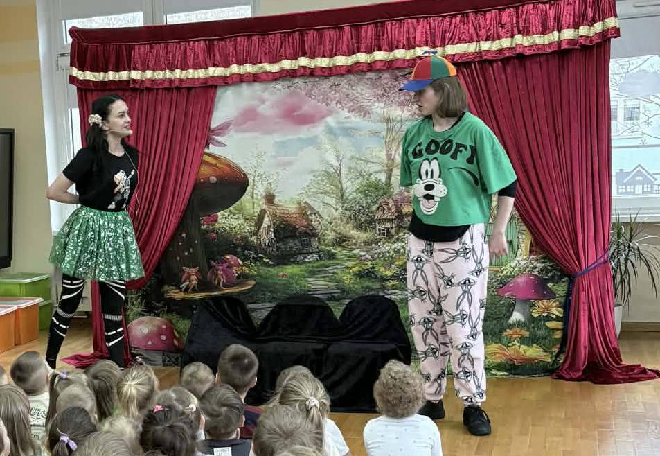 Dzieci siedzą w sali na dywanie i ogladają przedstawienie w wykonaniu dwóch aktorów.