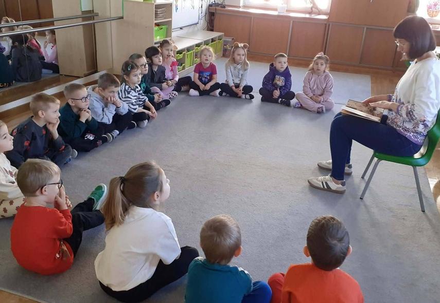 Mama jednego z dzieci siedzi na krześle i czyta dzieciom bajkę.