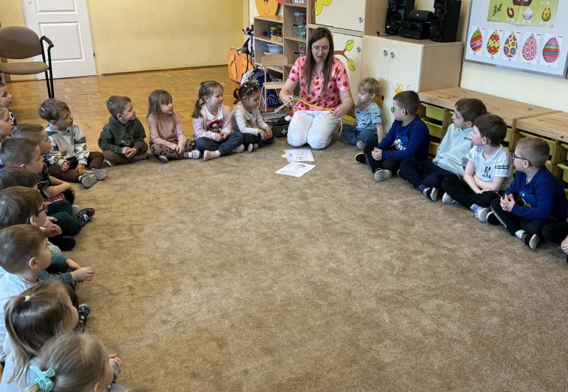 Dzieci siedzą w kole na dywanie z panią pielęgniarką.