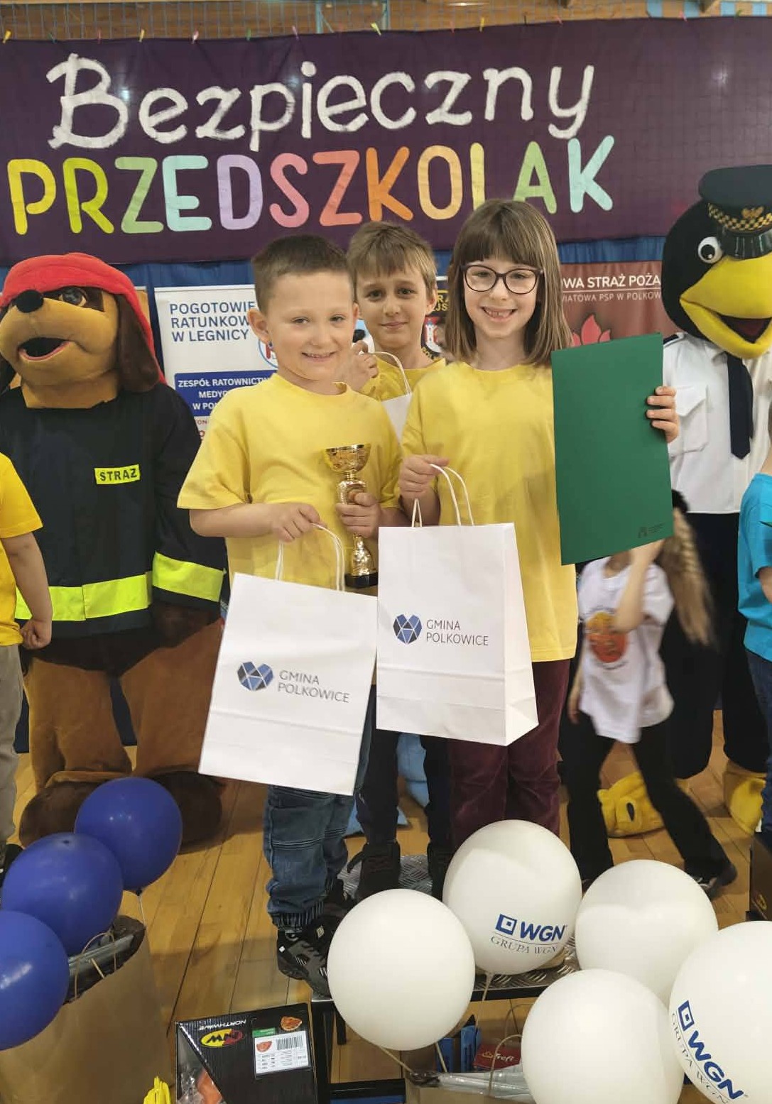 Troje dzieci ubranych w ż&oacute;łte koszulki i trzymając w ręku dyplomy stoi na podium.