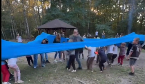 Dzieci w grupie otoczone są niebiską gumą i pr&oacute;bują ją rozciągnąć.