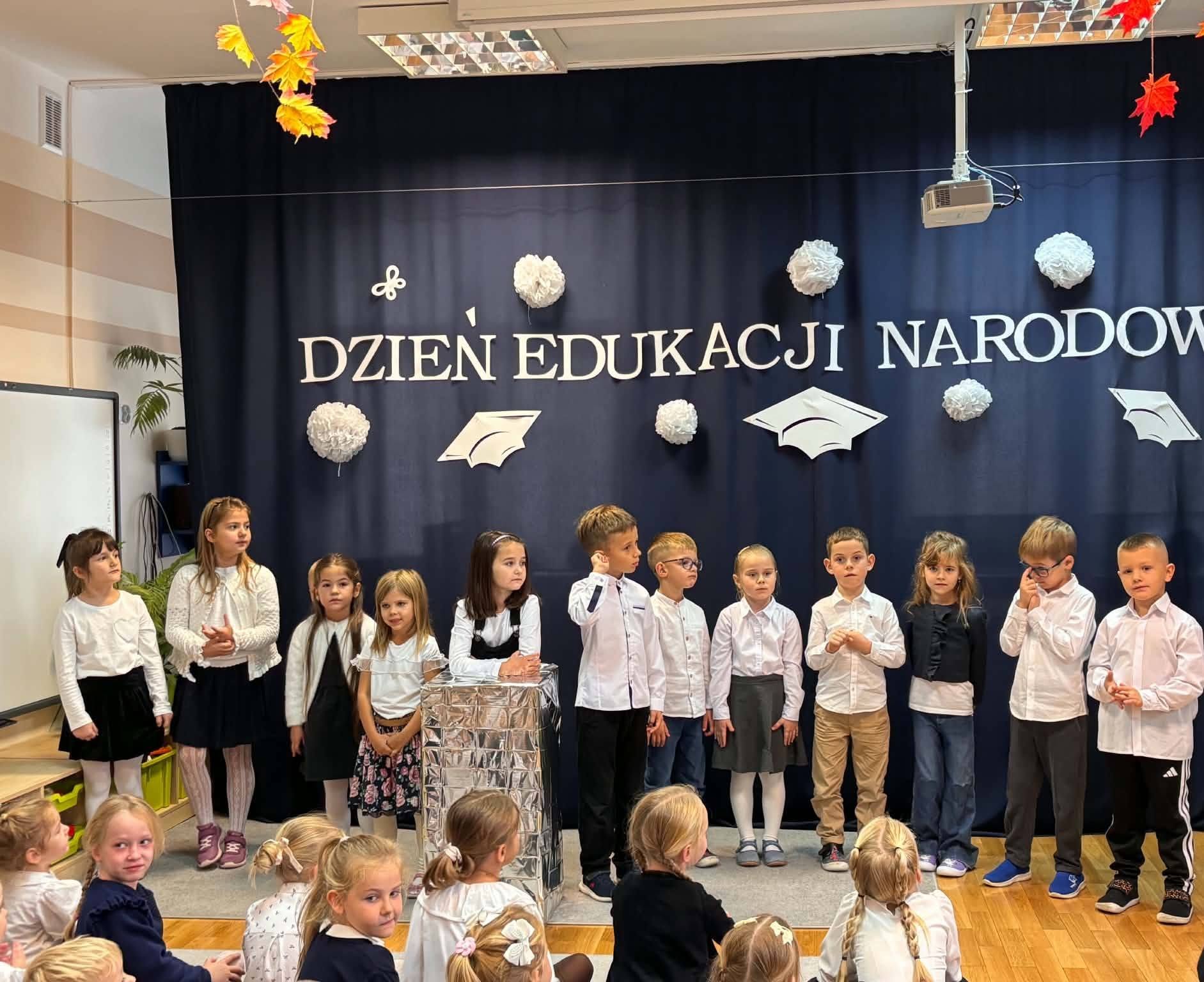 Dzieci ubrane na galowo stoją w p&oacute;łkolu na tle napisu dzień edukacji narodowej i recytują wierszyki.