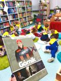 Dzieci siedzą na małych dywanikach w bibliotece i słuchają pani bibliotekarki.