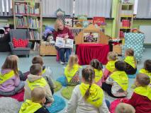 Dzieci siedzą na małych dywanikach w bibliotece i słuchają pani bibliotekarki.