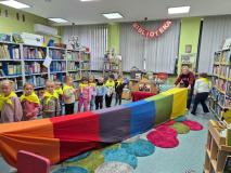 Dzieci siedzą na małych dywanikach w bibliotece i słuchają pani bibliotekarki.