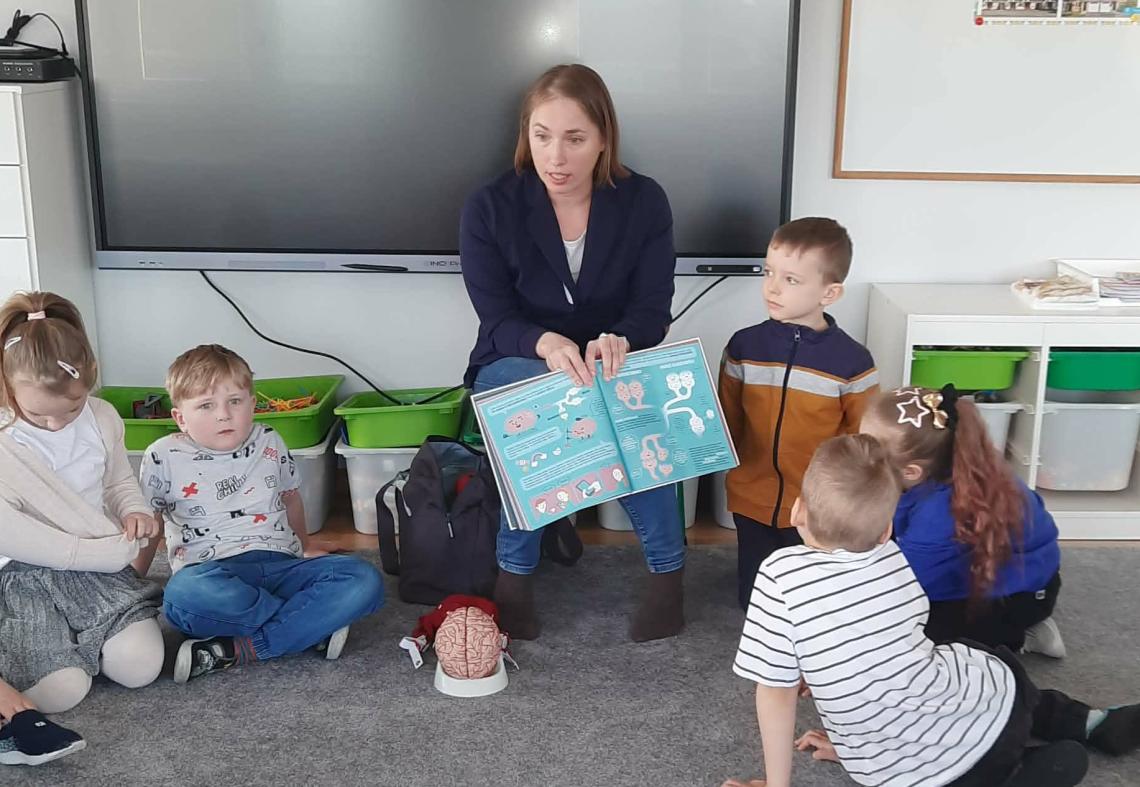 Mama dziecka trzyma w ręku książkę , a na dywanie leży makieta mózgu. Dzieci siedzą w kole na dywanie.