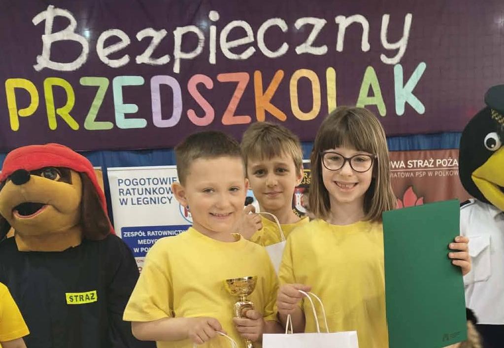 Troje dzieci ubranych w żółte koszulki i trzymając w ręku dyplomy stoi na podium.