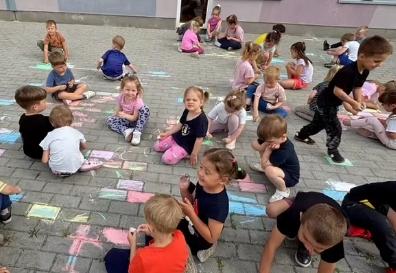 Grupa dzieci rysuje kredą na tarasie  przedszkola.